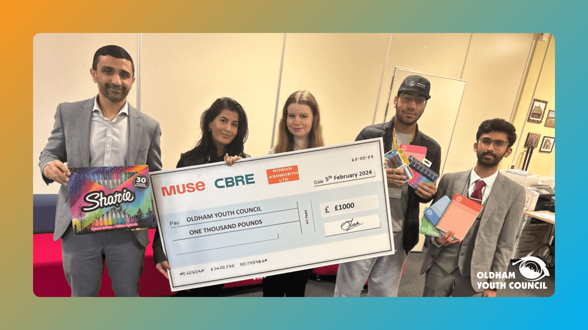 (L-R) Councillor Shaid Mushtaq, Leah, Adriana, Adam & Prem. Shaid Mushtaq holding a box of sharpies, Leah and Adriana holding a large cheque paid to the Oldham Youth Council for £1,000, Adam holding highlighters and markers, and Prem holding notebooks. The image is on a background of a gradient from orange to blue, with the Oldham Youth Council logo in the bottom right.